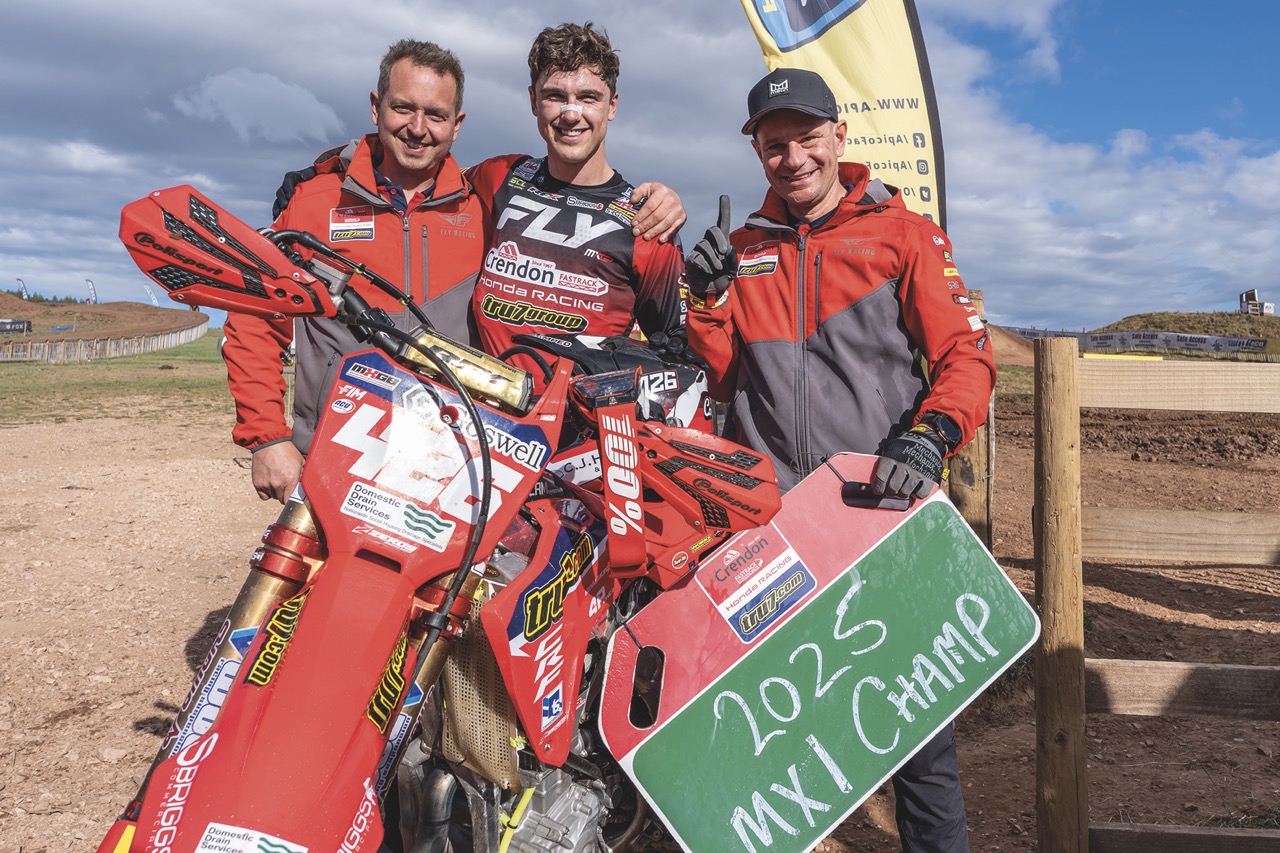 A group of three men wearing red and standing in front of a red motorbike on a dirt road