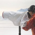 A person in an orange coat looking at a camera under a sheet as it snows around them