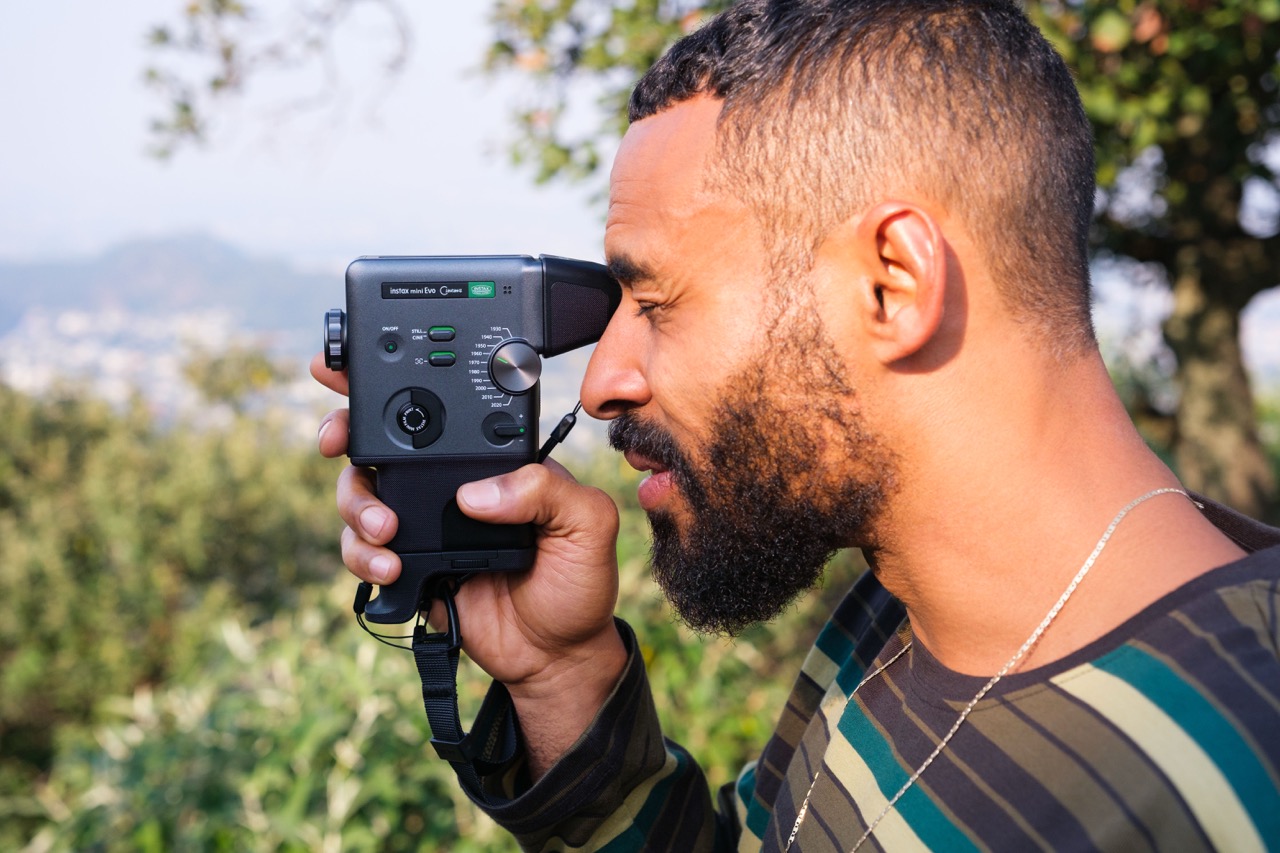 A man holding a vintage camcorder up to his eye, standing outside in a green area