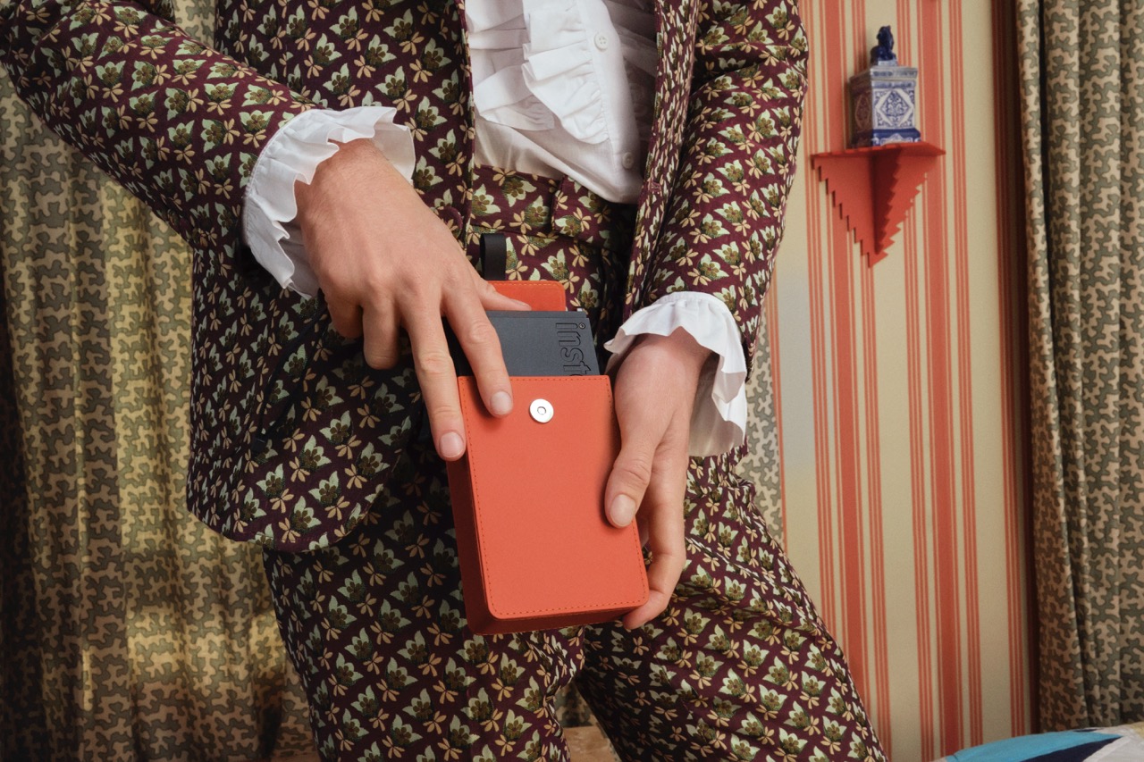 A person wearing a patterned suit, placing an instax printer in a bright orange case