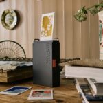 A black and orange instax printer with a photo coming from the top and photos spread on the table around it