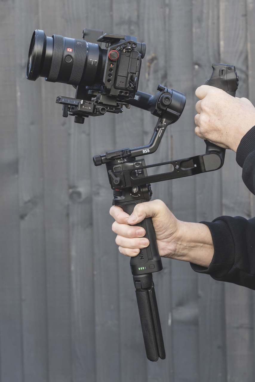 Hands holding a DJI gimbal vertically, against a wooden fence