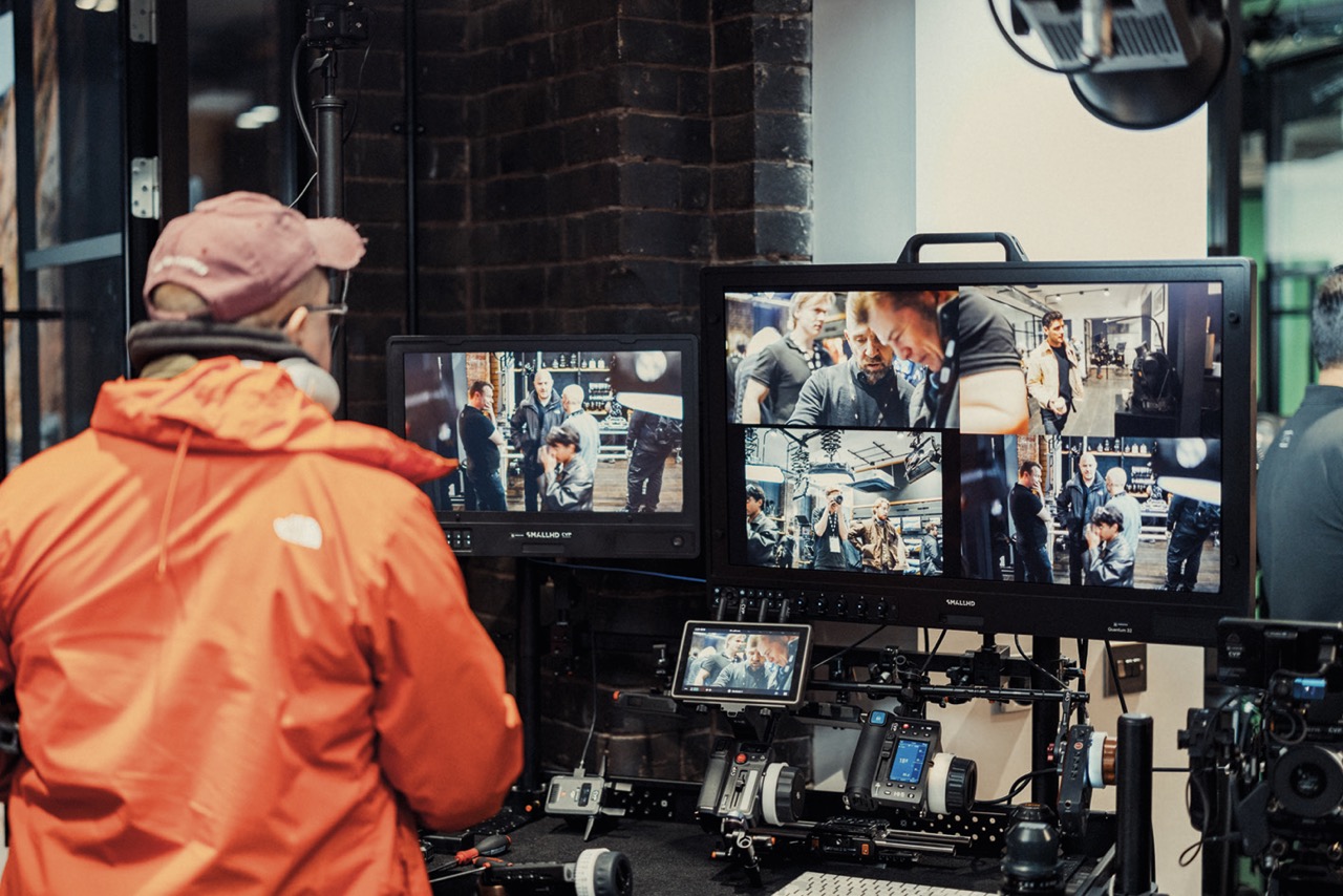 A person wearing a cap and orange coat staring at multiple screens in a studio