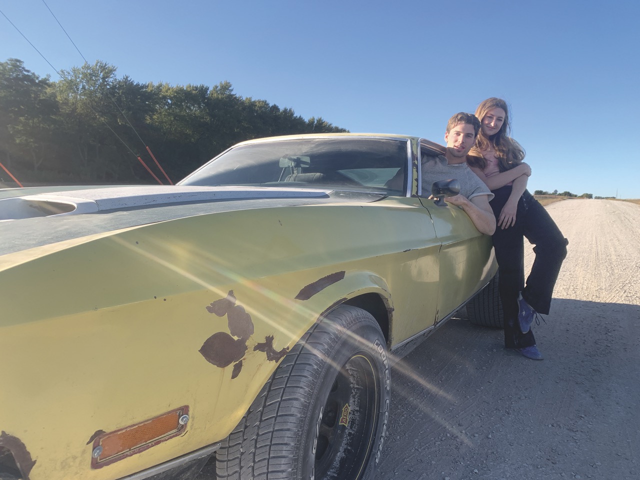 A girl leaning on a vintage yellow car and a boy leaning out of the window