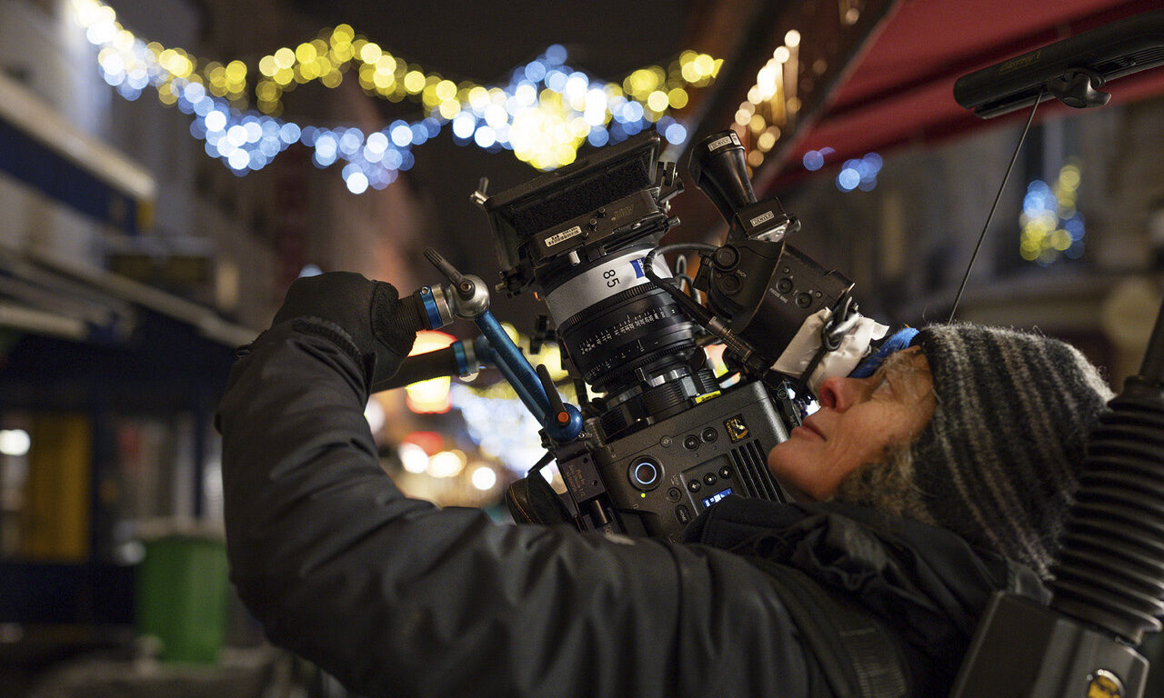 A person standing in a street at night, leaning back while holding a large, professional camera with a Zeiss lens attached