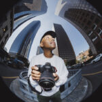A distorted fisheye image of a person holding a camera in a tall cityscape