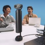 Two people smiling as they sit with laptops at the end of a table with a webcam and laptop at the other end. They sit in front of a light blue background