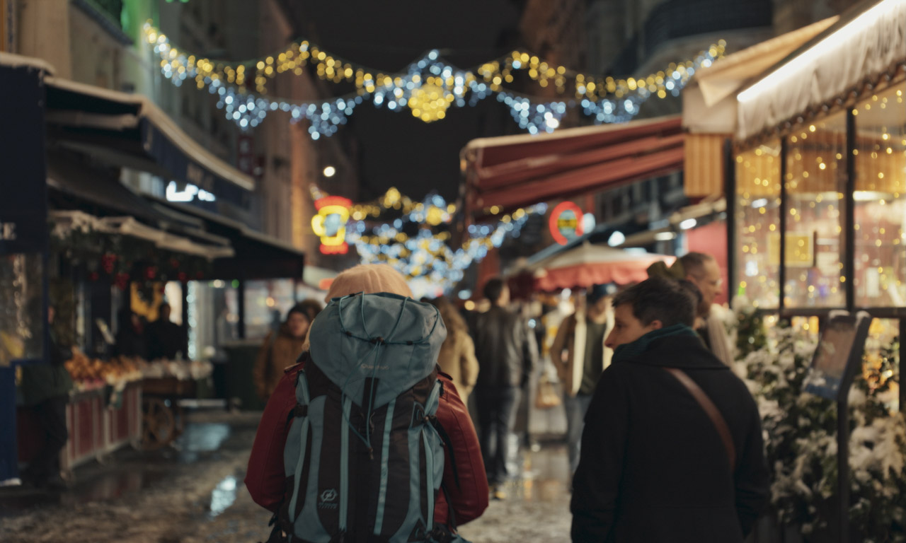 A busy night street with lights and open shops. A person with a backpack walks through the centre